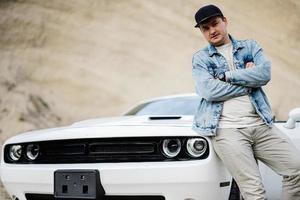 Handsome man in jeans jacket and cap is standing near his white muscle car in career. photo