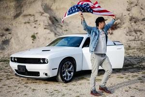 Handsome man in jeans jacket and cap with USA flag near his white american muscle car in career. photo
