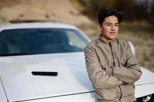 Teenager boy stand near white muscle car. Young teen driver. photo