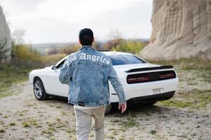 Back of man in jeans jacket and cap walking  near his white muscle car in career. Los Angeles. photo