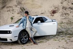 Handsome man in jeans jacket and cap is standing near his white muscle car in career and speaking on phone. photo