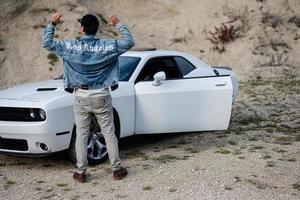 la parte posterior del hombre con chaqueta de jeans y gorra está de pie, cerca de su auto deportivo blanco en carrera. los Angeles. foto