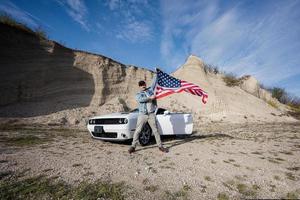 Handsome man in jeans jacket and cap with USA flag near his white american muscle car in career. photo