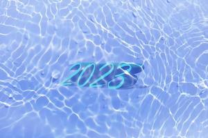 The numbers 2023 lying on water in swimming pool. Defocus blurred transparent blue colored clear calm water surface texture with splashes and bubbles. Trendy abstract nature background. New year 2023. photo