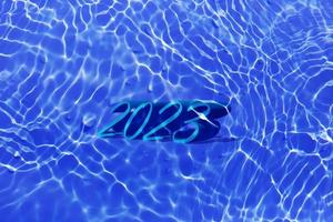 The numbers 2023 lying on water in swimming pool. Defocus blurred transparent blue colored clear calm water surface texture with splashes and bubbles. Trendy abstract nature background. New year 2023. photo