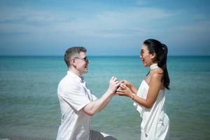 un joven blanco que pide matrimonio a una chica asiática con un anillo de diamantes en una playa foto