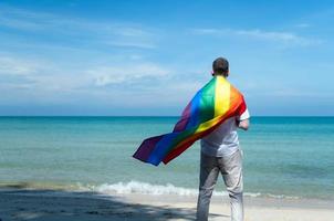 joven gay sosteniendo banderas del arco iris mientras está de pie en la playa foto