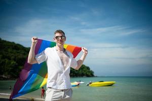 Young gay man holding rainbow flags while standing on the beach photo