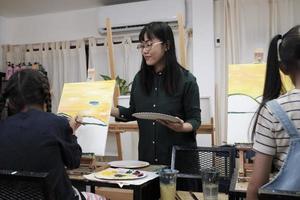 A female Asian teacher teaches and demonstrates to the children on acrylic color picture painting on canvas in art classroom, creatively learning with skill at the elementary school studio education. photo