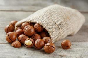 Macadamia nuts spilled out of the bag on a wooden background close-up with one peeled nut photo