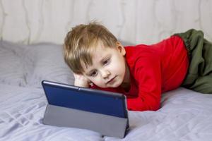 A cute boy is on the bed looking at a tablet. Online training, communication on the Internet. Funny smiling child. photo