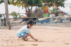 Asian cute little girl playing or making sand castle or digging with sand on tropical beach. Children with beautiful sea, sand and blue sky. Happy kids on vacations at seaside running on the beach. photo