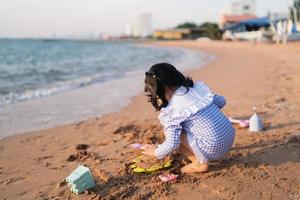 Asian cute little girl playing or making sand castle or digging with sand on tropical beach. Children with beautiful sea, sand and blue sky. Happy kids on vacations at seaside running on the beach. photo