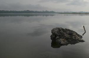 Front view of a large rock in Karangkates lake Indonesia with calm water conditions at sunrise with a mountains background and a foggy sky photo