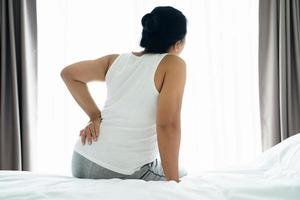 Adult Asian woman is sitting on the bed and holding her lower back suffering from injured back. Health care and back pain concept.