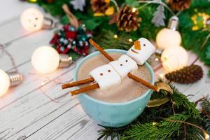 Big mug with hot Cappuccino with melted marshmallow snowman on a background of bokeh Christmas garlands and christmas tree. photo
