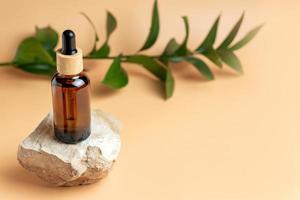 Amber glass bottle of essential oil or serum with pipette on stone podium with and green branch on a beige background. Natural skincare cosmetic concept photo