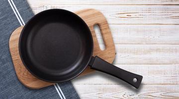 Black fry pan and board with napkin on wooden table. Top view mockup photo