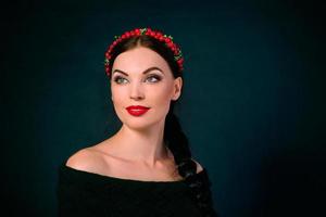 portrait of smiling young beautiful brunette with red wreath and braid on dark background photo