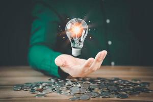 Man holding a glowing abstract light bulb with pile money cions on a wooden table, business problem solving ideas and creative marketing ideas Inventing and learning new things. photo