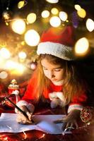 Little girl in a Santa hat and red dress under Christmas tree is dreaming, waiting for the holiday, lying on a plaid blanket. A letter on piece of paper, gifts. New year, Christmas. defocus lights photo