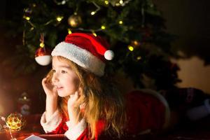 Little girl in a Santa hat and red dress under Christmas tree is dreaming, waiting for the holiday, lying on a plaid blanket. A letter on piece of paper, gifts. New year, Christmas. defocus lights photo