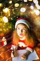 Little girl in a Santa hat and red dress under Christmas tree is dreaming, waiting for the holiday, lying on a plaid blanket. A letter on piece of paper, gifts. New year, Christmas. defocus lights photo