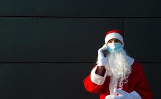 santa claus con una máscara médica con un teléfono inteligente. saludos en línea, servicios de pedidos para navidad y año nuevo. distancia social en la epidemia de coronavirus, una nueva realidad. copie el espacio foto