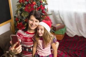 mamá e hija están sentadas cerca del árbol de navidad, abrazándose, tomando fotos y selfies por teléfono, comunicándose por videocomunicación, divirtiéndose. navidad, fin de año en casa. copie el espacio