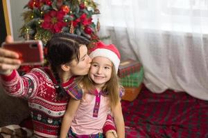 Mom and daughter are sitting near the Christmas tree, hugging, taking photos and selfies on the phone, communicating via video communication, having fun. Christmas, new year's eve at home. Copy space