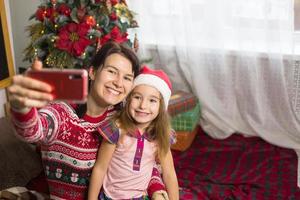 Mom and daughter are sitting near the Christmas tree, hugging, taking photos and selfies on the phone, communicating via video communication, having fun. Christmas, new year's eve at home. Copy space