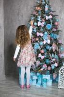 A little girl reaches for a Christmas tree toy with her hand. Christmas decor, waiting for a holiday and a miracle. New Year. White, pink and blue decor, Golden lights garlands photo