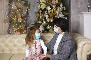 abuela y nieta en el sofá de la sala de estar con decoración navideña abrazándose con máscaras médicas en la cara. unas vacaciones familiares durante el brote de coronavirus y enfermedad. Año Nuevo foto