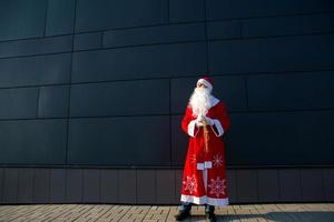 un joven vestido como santa en la calle, parado contra una pared moderna gris. barba blanca, traje rojo con sombrero. navidad, año nuevo. helada del abuelo ruso. fondo moderno gris, espacios de copia foto