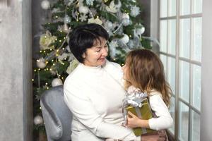 Grandmother with a little girl on the background of Christmas decorations and a large window. Family holiday, emotions, gift box. Granddaughter on granny's lap. New Year photo