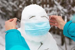 muñeco de nieve con una máscara médica: una nueva realidad, protección contra enfermedades, infecciones, vida en la epidemia de covid. las manos de las mujeres se ponen una máscara de muñeco de nieve. actividades al aire libre de la familia de invierno foto