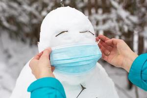 muñeco de nieve con una máscara médica: una nueva realidad, protección contra enfermedades, infecciones, vida en la epidemia de covid. las manos de las mujeres se ponen una máscara de muñeco de nieve. actividades al aire libre de la familia de invierno foto