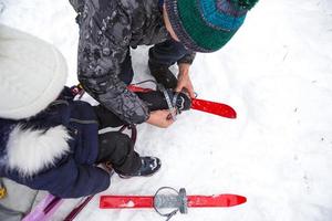 Dad helps his little daughter to put on children's plastic skis. Sports training, skiing, support and assistance. Winter active sports outdoor since childhood