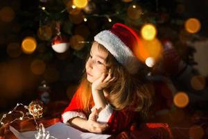 la niña pequeña con un sombrero de santa y un vestido rojo bajo el árbol de navidad está soñando, esperando las vacaciones, acostada sobre una manta a cuadros. una carta en papel, regalos. año nuevo, navidad. luces de desenfoque foto