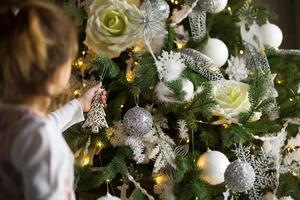 A little girl reaches for a Christmas tree toy with her hand. Christmas decor, waiting for a holiday and a miracle. New Year. White, pink and blue decor, Golden lights garlands photo