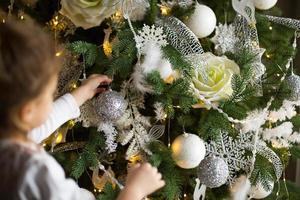 A little girl reaches for a Christmas tree toy with her hand. Christmas decor, waiting for a holiday and a miracle. New Year. White, pink and blue decor, Golden lights garlands photo