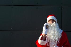 moderno santa claus hablando por teléfono. solicitando los servicios de un animador para navidad y año nuevo. saludos en línea a través de Internet y comunicación móvil, distancia social foto