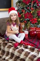 A little girl holding a Teddy bear, sitting on a plaid blanket in the Christmas decorations near a Christmas tree with boxes of gifts and a Santa hat. New year, children's game photo