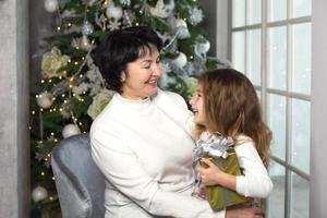 Grandmother with a little girl on the background of Christmas decorations and a large window. Family holiday, emotions, gift box. Granddaughter on granny's lap. New Year photo