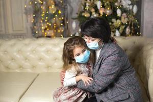 Grandmother and granddaughter on the sofa in the living room with Christmas decor hugging in medical masks on their faces. A family holiday during the outbreak of coronavirus and disease. New Year photo