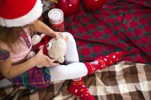 A little girl holding a Teddy bear, sitting on a plaid blanket in the Christmas decorations near a Christmas tree with boxes of gifts and a Santa hat. New year, children's game photo