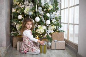 niña en hermoso vestido está sentada bajo el árbol de navidad con caja de regalo y lazo. luz de un ventanal, cuarto oscuro, confort hogareño, navidad, magia y alegría. año nuevo, decoración blanca foto