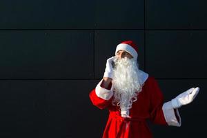 moderno santa claus hablando por teléfono. solicitando los servicios de un animador para navidad y año nuevo. saludos en línea a través de Internet y comunicación móvil, distancia social foto