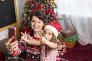 Mom and daughter are sitting near the Christmas tree, hugging, taking photos and selfies on the phone, communicating via video communication, having fun. Christmas, new year's eve at home. Copy space