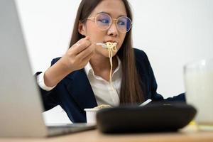 mujer de negocios ocupada y cansada comiendo espagueti para el almuerzo en la oficina y trabajando para entregar estados financieros a un jefe. con exceso de trabajo y poco saludable para comidas preparadas, concepto de agotamiento. foto